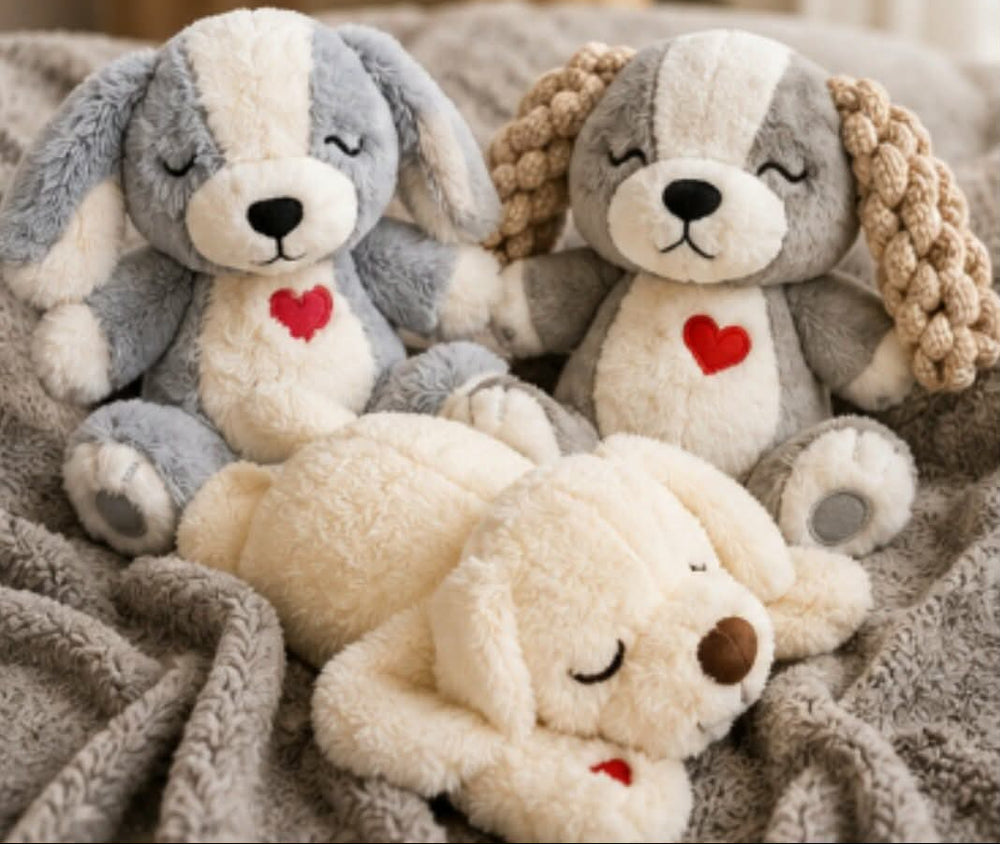 Three Hug Me Dogs plush dog toys arranged together on a soft gray blanket in a cozy indoor setting. Two dogs are sitting upright and one is lying down, all in cream and gray tones with small red hearts on their chests.